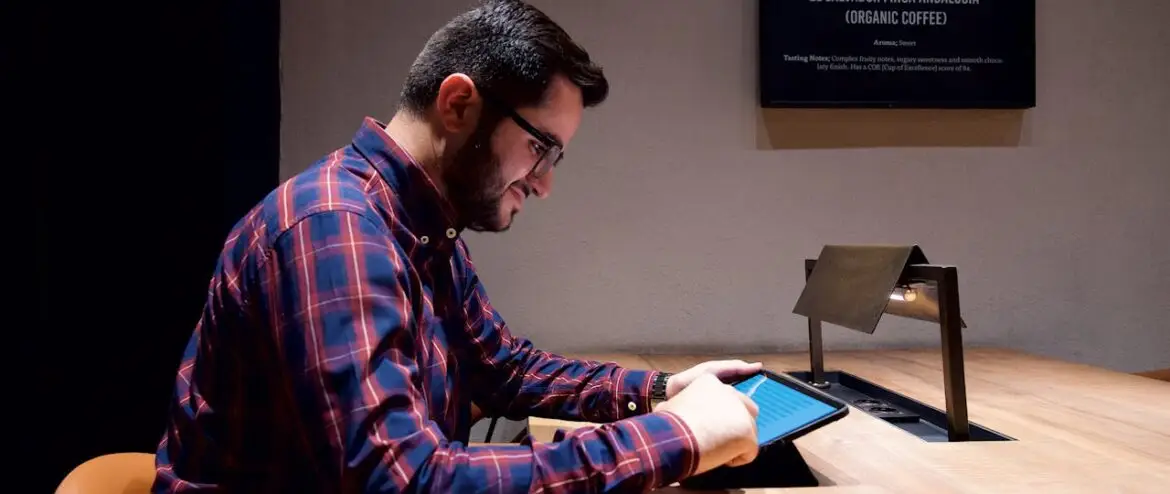 A man interacts with a touchscreen tablet at a stylish wooden desk in a cozy coffee shop.