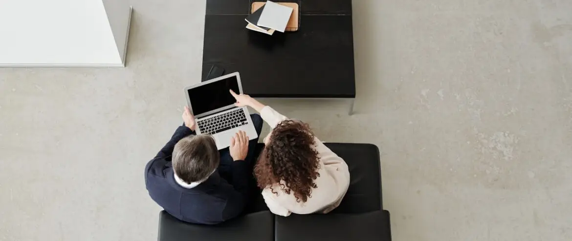 Two colleagues collaborating on a laptop from a top view, seated on a leather sofa.